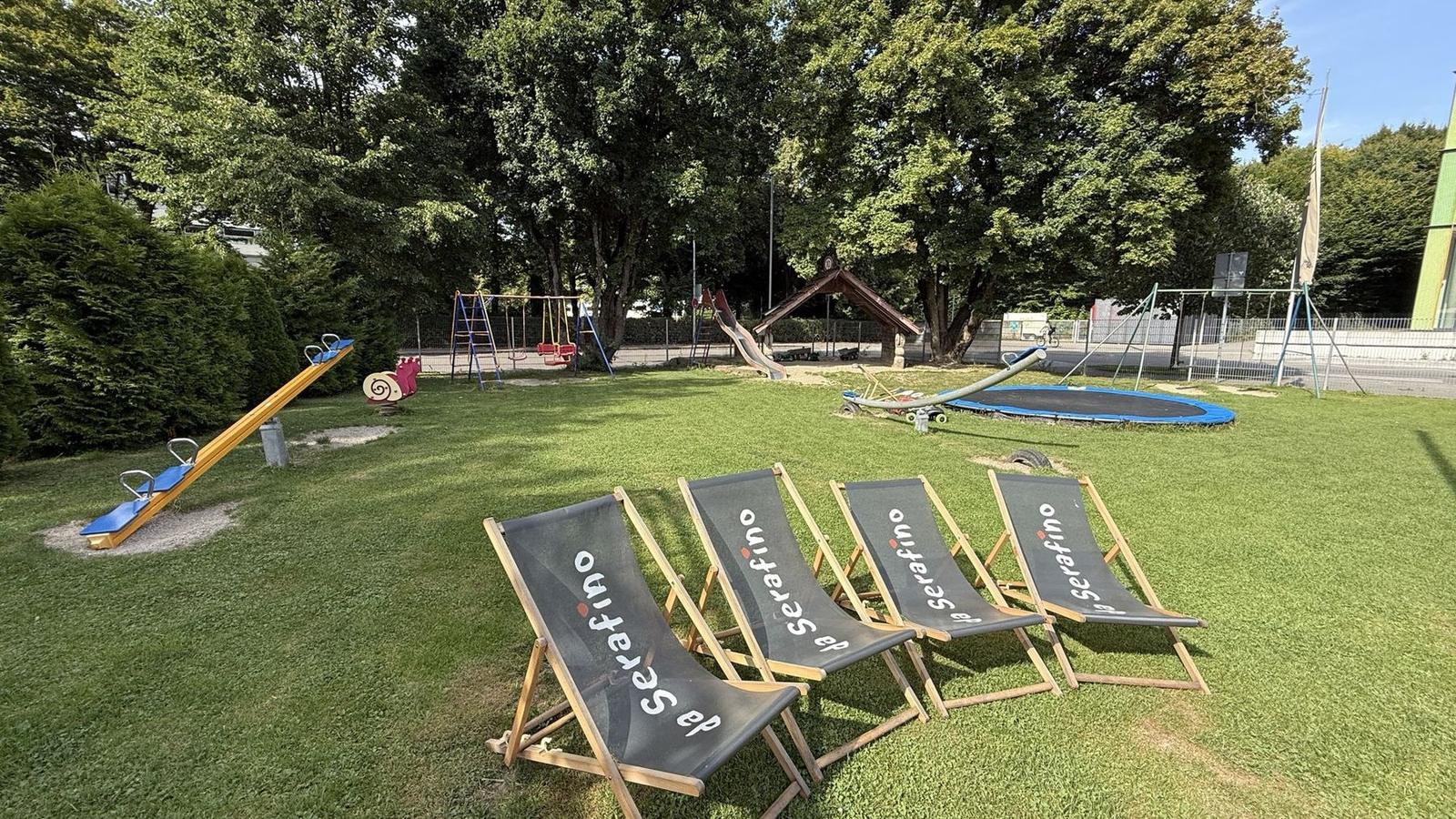 Spielplatz im Grünen mit Wippe, Schaukeln und Trampolin, davor vier Liegestühle mit da-Serafino-Schriftzug