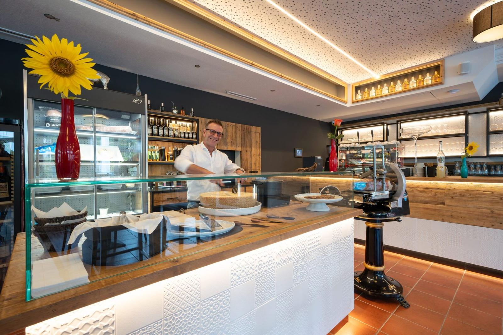Der Gastgeber lächelnd hinter der Theke mit Parmesanlaib, im Hintergrund die beleuchtete Bar